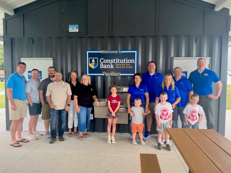 Group of adults and children posing together in front of a blue Constitution Bank Pavilion sign at an outdoor pavilion.
