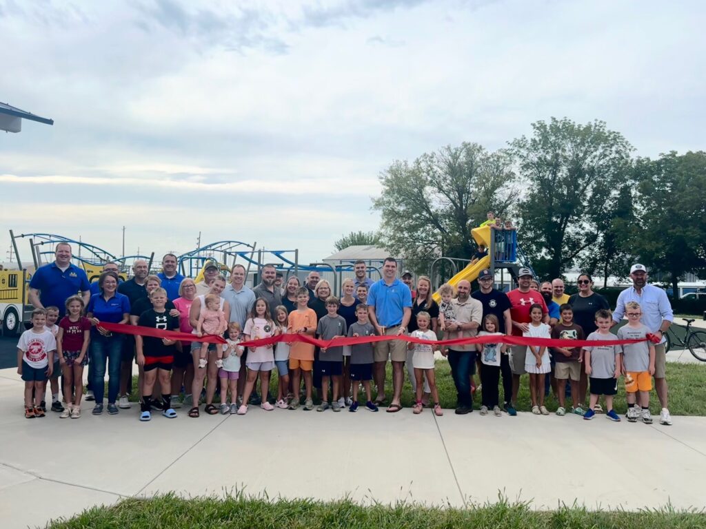 Large group of adults and children posing behind a red ribbon at a playground ribbon-cutting ceremony, with blue playground equipment and trees in the background.